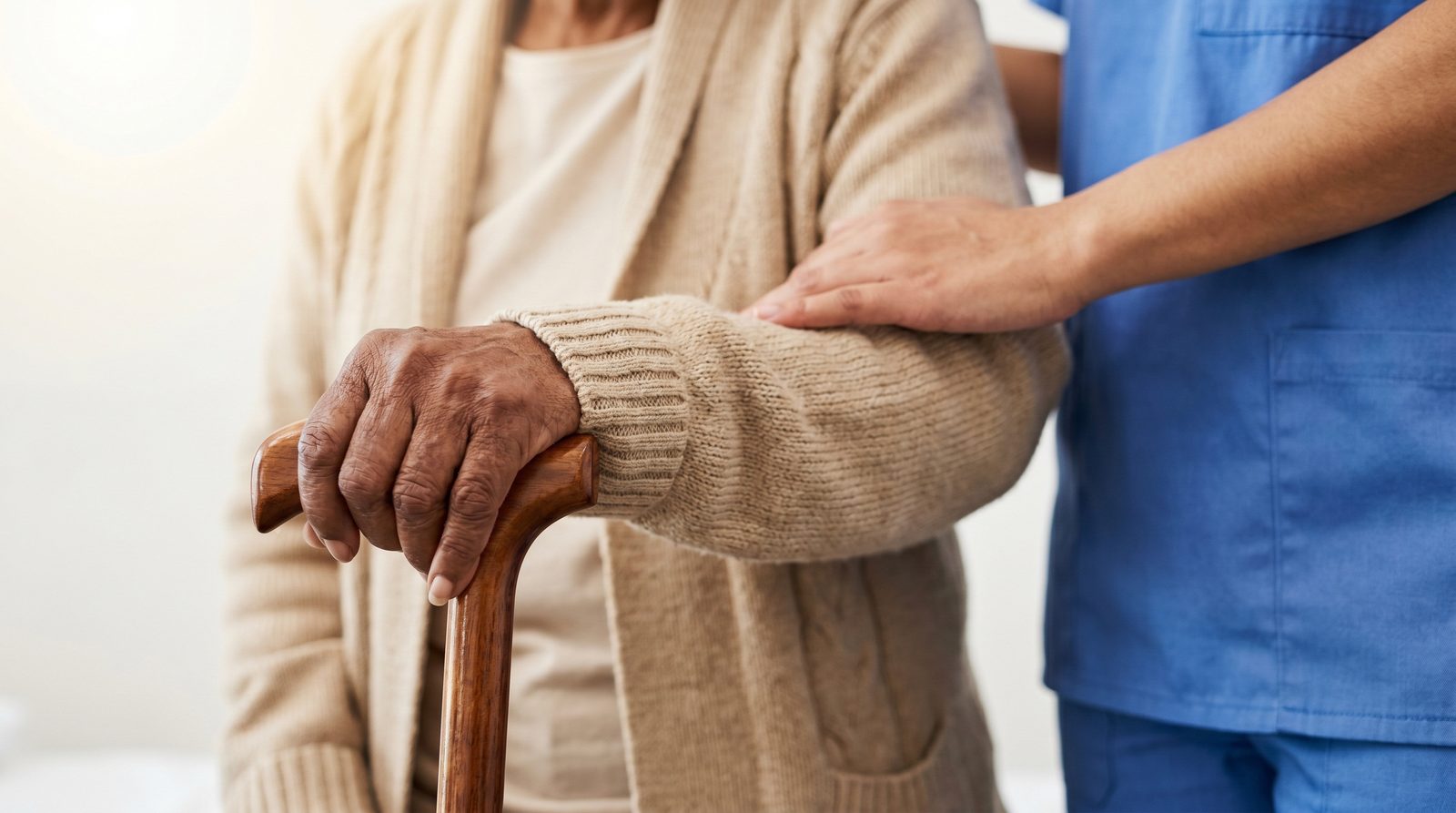 A care worker's hand gently supporting a senior who is using a walking cane